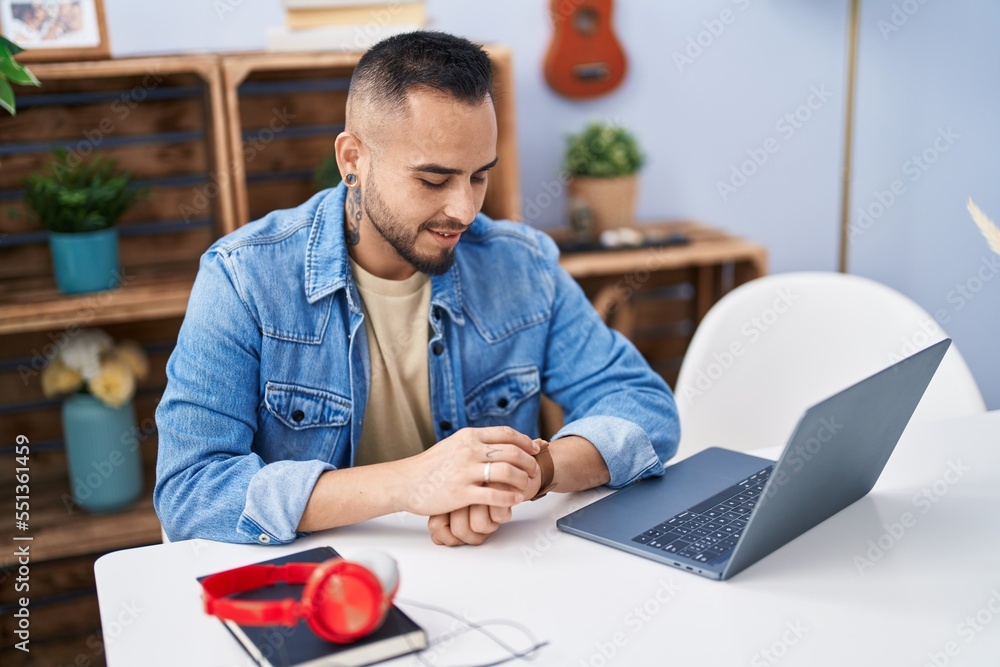 Fototapeta premium Young hispanic man sitting on table looking watch at home