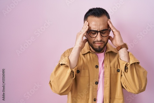 Young hispanic man standing over pink background with hand on head for pain in head because stress. suffering migraine.