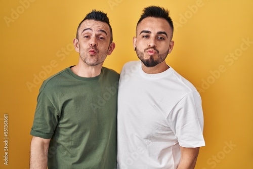Homosexual couple standing over yellow background looking at the camera blowing a kiss on air being lovely and sexy. love expression.