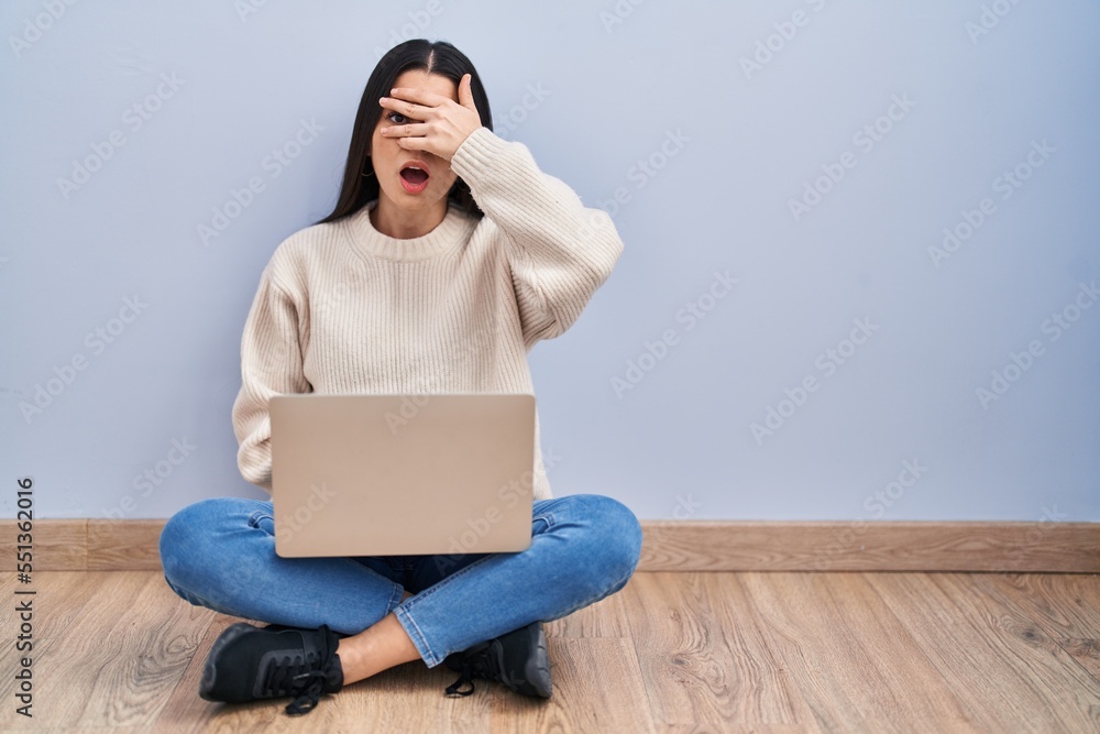 Young woman using laptop sitting on the floor at home peeking in shock covering face and eyes with hand, looking through fingers with embarrassed expression. Young woman using laptop sitting on the floor at home peeking in shock covering face and eyes with hand, looking through fingers with embarrassed expression.