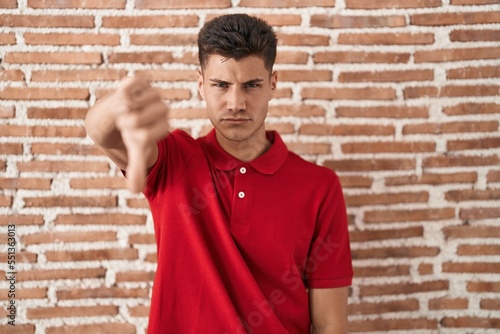 Young hispanic man standing over bricks wall looking unhappy and angry showing rejection and negative with thumbs down gesture. bad expression.