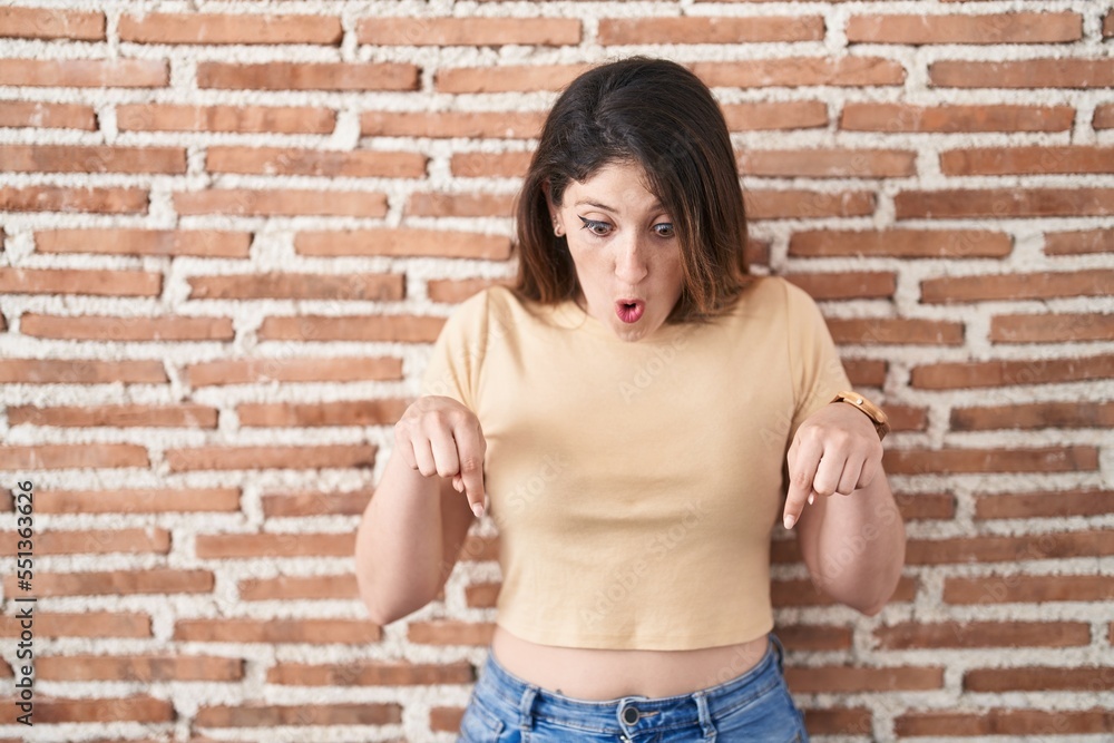 Young brunette woman standing over bricks wall pointing down with fingers showing advertisement, surprised face and open mouth
