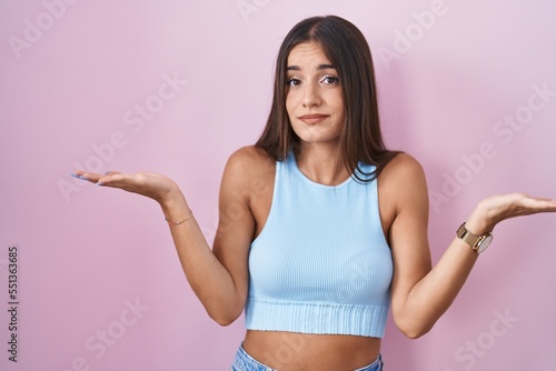 Young brunette woman standing over pink background clueless and confused expression with arms and hands raised. doubt concept.