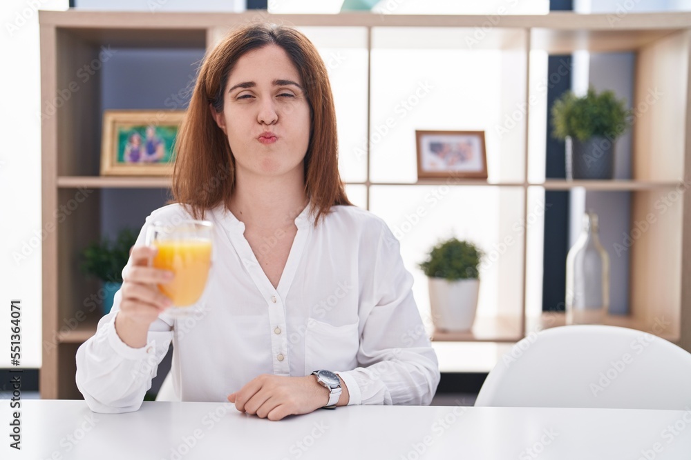 Brunette woman drinking glass of orange juice puffing cheeks with funny face. mouth inflated with air, crazy expression. Brunette woman drinking glass of orange juice puffing cheeks with funny face. mouth inflated with air, crazy expression.