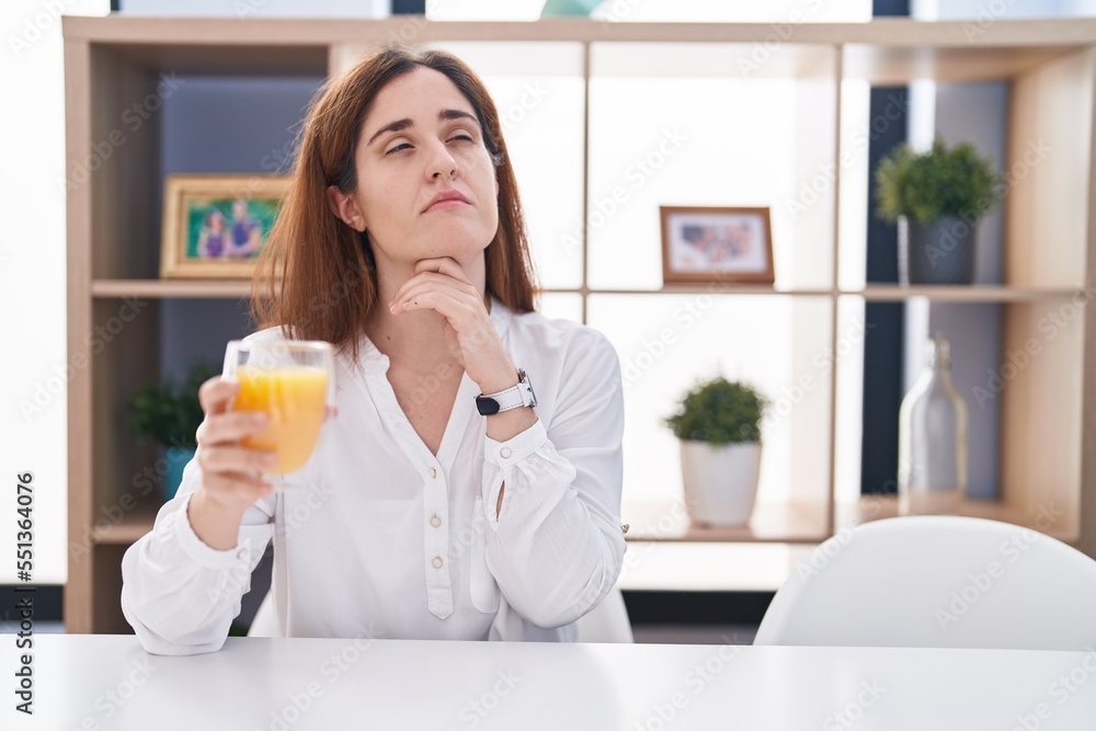 Brunette woman drinking glass of orange juice with hand on chin thinking about question, pensive expression. smiling with thoughtful face. doubt concept. Brunette woman drinking glass of orange juice with hand on chin thinking about question, pensive expression. smiling with thoughtful face. doubt concept.