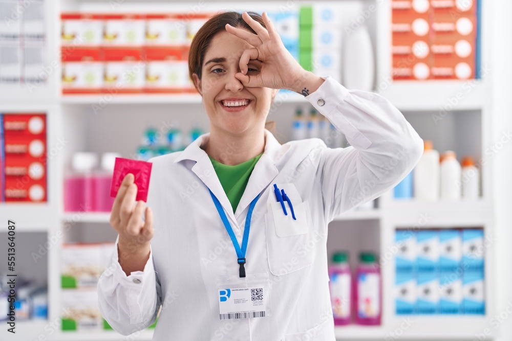 Brunette woman working at pharmacy drugstore holding condom smiling happy doing ok sign with hand on eye looking through fingers Brunette woman working at pharmacy drugstore holding condom smiling happy doing ok sign with hand on eye looking through fingers