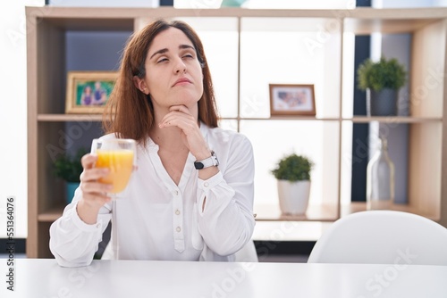 Brunette woman drinking glass of orange juice with hand on chin thinking about question  pensive expression. smiling with thoughtful face. doubt concept.