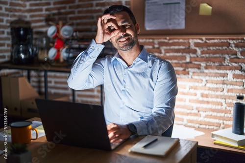 Hispanic man with beard working at the office at night doing ok gesture with hand smiling, eye looking through fingers with happy face.