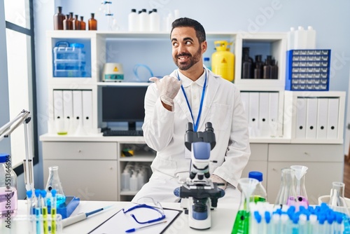 Young hispanic man with beard working at scientist laboratory smiling with happy face looking and pointing to the side with thumb up.