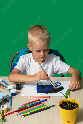 Boy draws with brush, flower in pot on table, paints, pencils, books, cubes. Selective focus. Learning to draw, hobbies in your free time.
