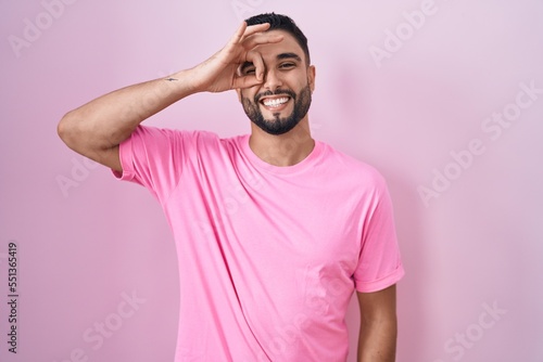 Hispanic young man standing over pink background doing ok gesture with hand smiling, eye looking through fingers with happy face.