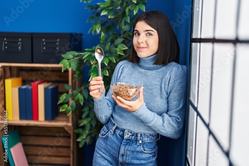 Young hispanic woman eating healthy whole grain cereals with spoon smiling looking to the side and staring away thinking.
