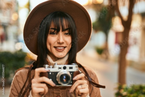 Brunette woman wearing winter hat smiling using vintage camera outdoors at the city