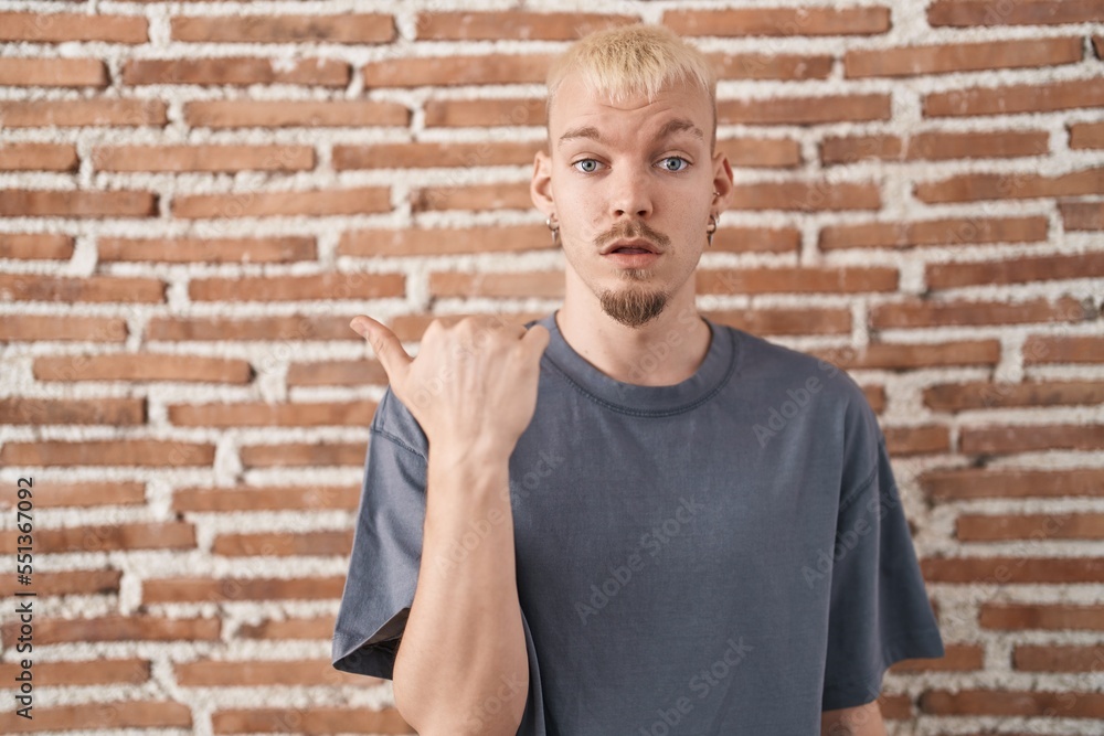 Young caucasian man standing over bricks wall surprised pointing with hand finger to the side, open mouth amazed expression. Young caucasian man standing over bricks wall surprised pointing with hand finger to the side, open mouth amazed expression.