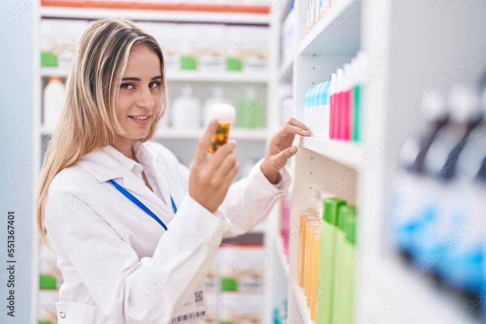 Young blonde woman pharmacist holding pills bottle of shelving at pharmacy