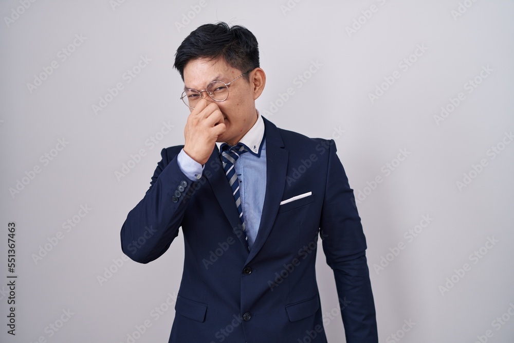 Young asian man wearing business suit and tie smelling something stinky and disgusting, intolerable smell, holding breath with fingers on nose. bad smell Young asian man wearing business suit and tie smelling something stinky and disgusting, intolerable smell, holding breath with fingers on nose. bad smell