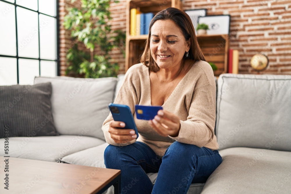 Fototapeta premium Middle age hispanic woman using smartphone and credit card sitting on sofa at home