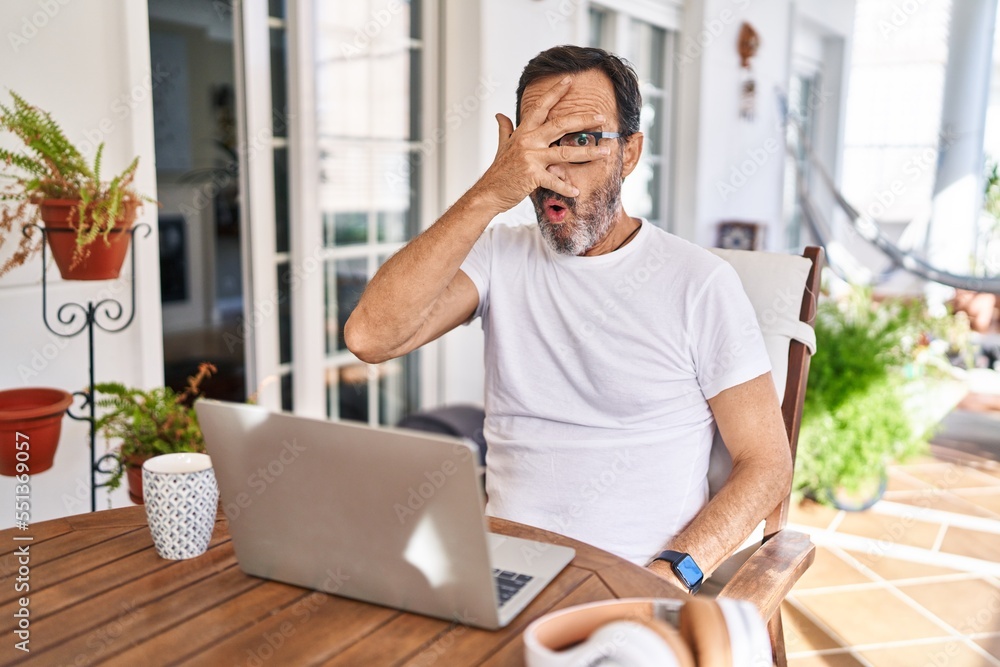 Middle age man using computer laptop at home peeking in shock covering face and eyes with hand, looking through fingers with embarrassed expression. Middle age man using computer laptop at home peeking in shock covering face and eyes with hand, looking through fingers with embarrassed expression.