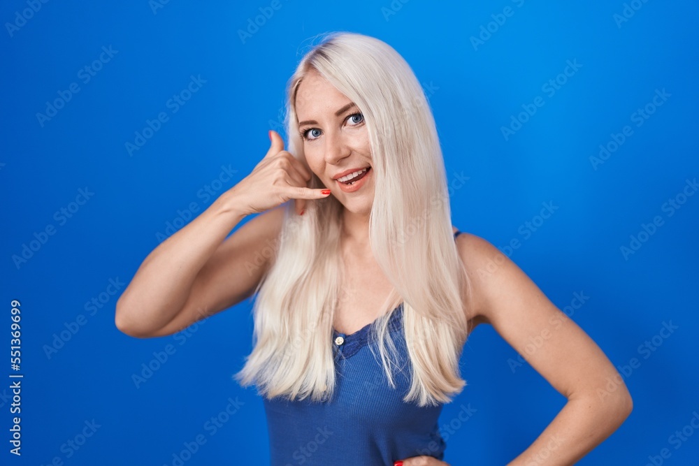 Caucasian woman standing over blue background smiling doing phone gesture with hand and fingers like talking on the telephone. communicating concepts. Caucasian woman standing over blue background smiling doing phone gesture with hand and fingers like talking on the telephone. communicating concepts.