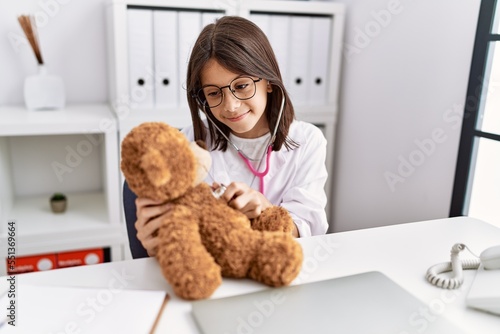 Young hispanic girl checking teddy bear health at doctor clinic