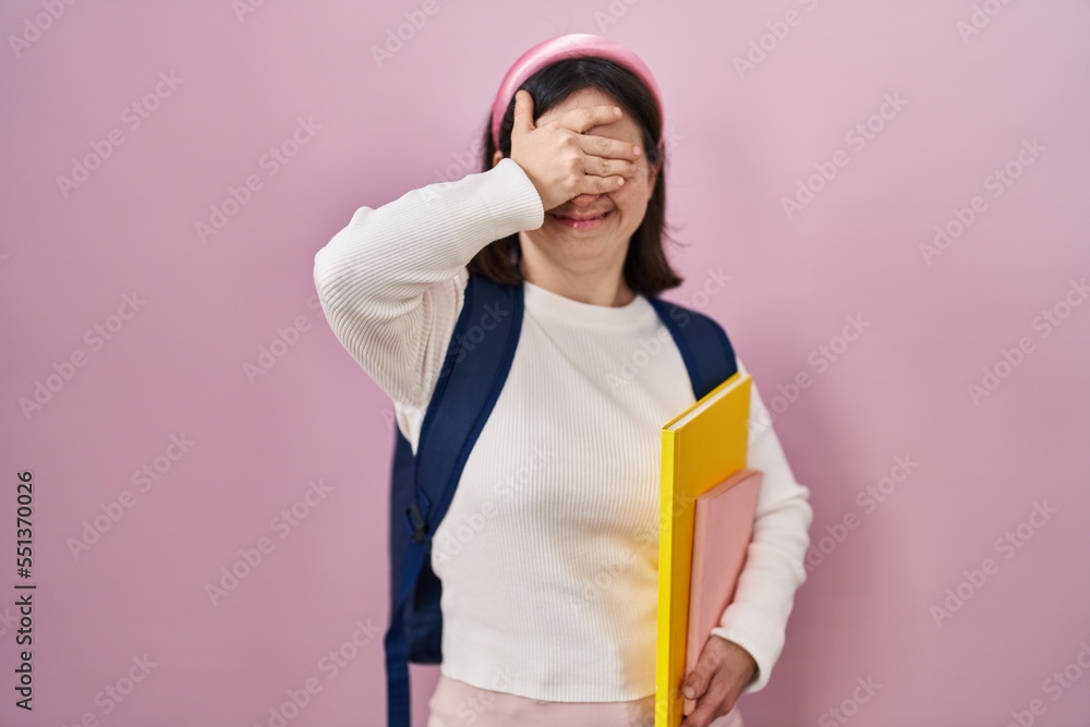Woman with down syndrome wearing student backpack and holding books smiling and laughing with hand on face covering eyes for surprise. blind concept. Woman with down syndrome wearing student backpack and holding books smiling and laughing with hand on face covering eyes for surprise. blind concept.
