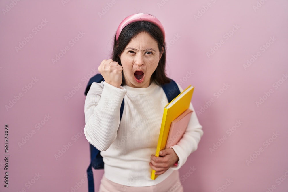 Woman with down syndrome wearing student backpack and holding books angry and mad raising fist frustrated and furious while shouting with anger. rage and aggressive concept. Woman with down syndrome wearing student backpack and holding books angry and mad raising fist frustrated and furious while shouting with anger. rage and aggressive concept.