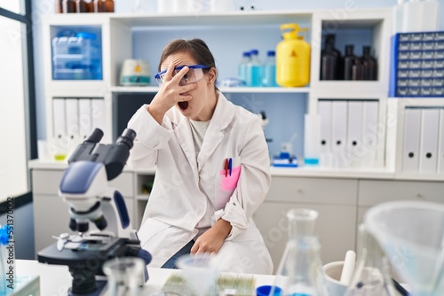 Hispanic girl with down syndrome working at scientist laboratory peeking in shock covering face and eyes with hand  looking through fingers with embarrassed expression.
