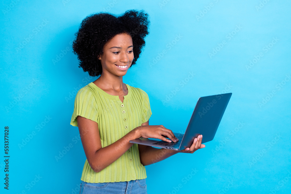 Photo of positive clever girl with perming coiffure dressed green t-shirt hold laptop doing homework isolated on blue color background Photo of positive clever girl with perming coiffure dressed green t-shirt hold laptop doing homework isolated on blue color background