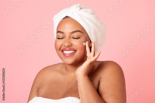 Happy black body positive woman applying beauty product on her face and smiling, lady wrapped in towel using face cream