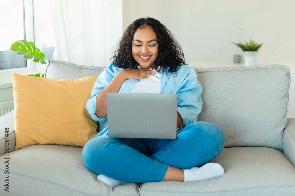 Happy black oversize lady using laptop at home, reading news or making video call with friends, resting on sofa at home Happy black oversize lady using laptop at home, reading news or making video call with friends, resting on sofa at home