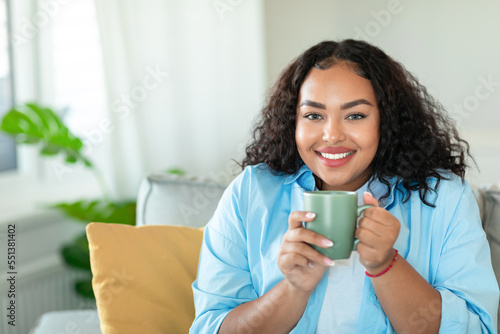 Domestic leisure. Happy black plus size lady drinking coffee at home  resting on comfortable sofa in cozy living room