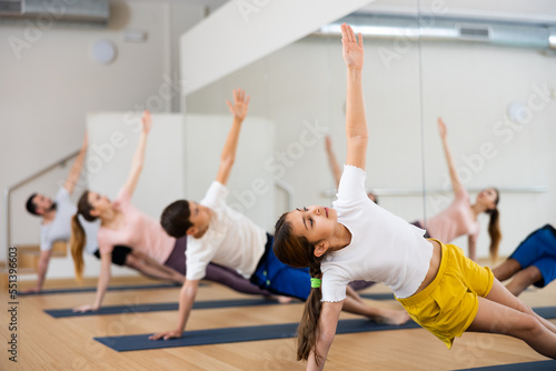 Girl exercising side plank on mat during family training in gym.