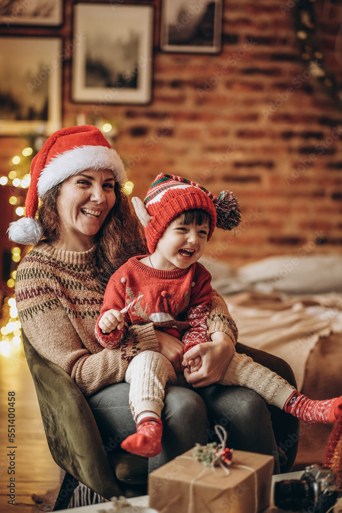 Obraz premium Happy baby with mother sit in armchair on christmas eve and kid eats christmas candy.