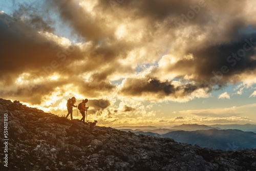 two hikers with their dog in silhouette descending a mountain at sunset on a cloudy day. couple doing trekking. sport and outdoor adventure.