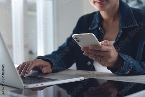 close up of young asian business woman using mobile smart phone and working on laptop computer with digital tablet on table at office.