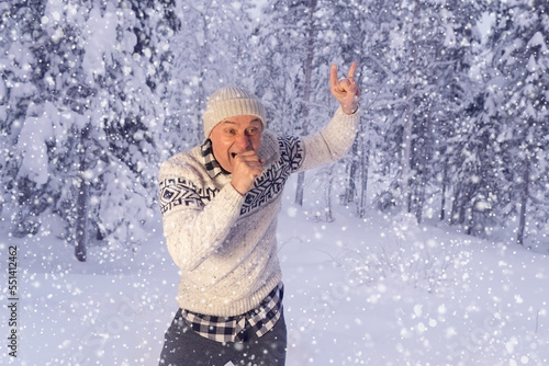 positive mature charismatic man 60 years in hat and Scandinavian sweater is merrily fooling around, imitates singing into microphone, grimacing in winter forest, concept of active lifestyle, good mood