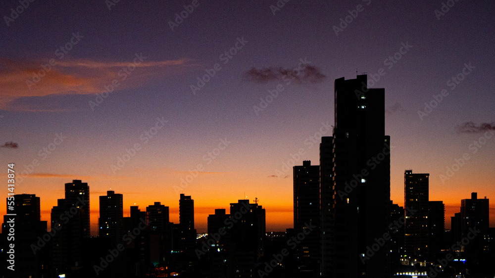 Urban sunset seen from Tirol, Natal, Rio Grande do Norte. Silhouettes of buildings formed by yellow-orange sunlight under a blue-purple sky.