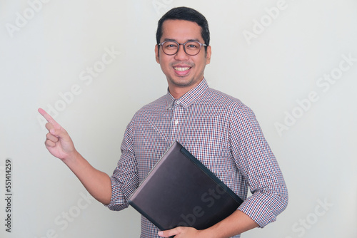 Adult Asian man smiling and pointing to the right side while holding laptop computer photo