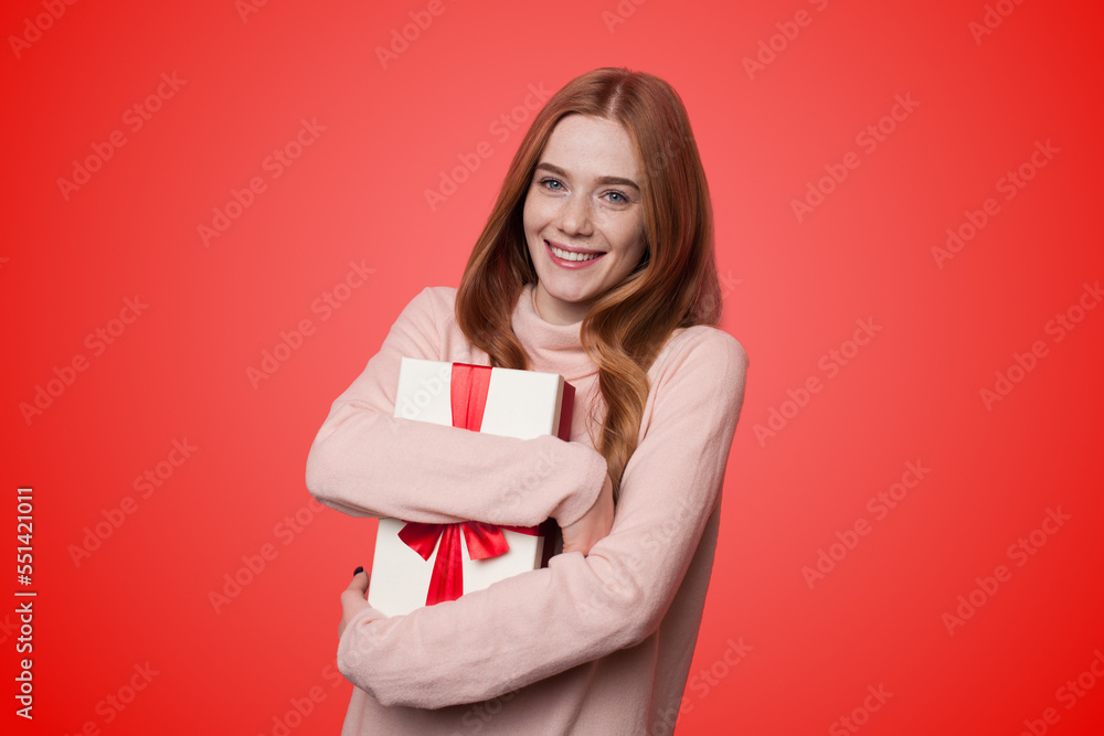 Photo of positive happy woman with red hair embracing gift box, looking and smiling at camera isolated on red color background. Christmas, or her birthday Photo of positive happy woman with red hair embracing gift box, looking and smiling at camera isolated on red color background. Christmas, or her birthday
