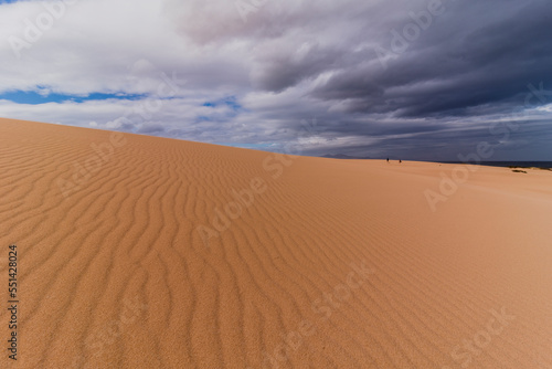 Sand dune elevation and sea shore with dark blue clouds and two people near ocean in distance 