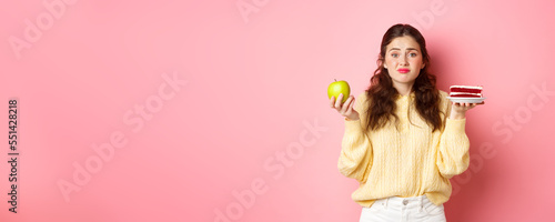 Young cute woman hesitating between delicious piece of cake dessert and green healthy apple, being on diet, standing indecisive against pink background