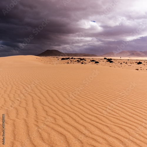 Structured sand dunes with dark volcano and highway against stormy and cloudy sky, Corralejo, Spain