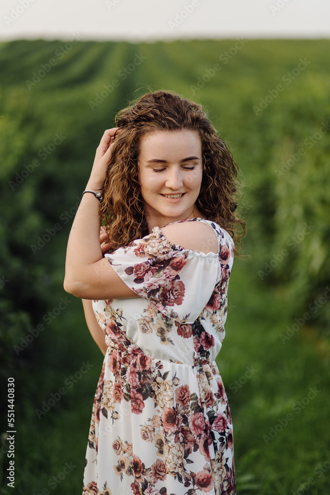 Happy young woman with brown curly hair, wearing a dress, posing outdoors in a garden Happy young woman with brown curly hair, wearing a dress, posing outdoors in a garden