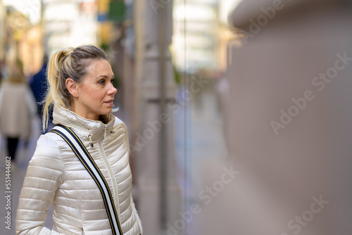 Gently smiling attractive middle-aged woman with blond hair and a ponytail looks interested through a shop window in a shopping street