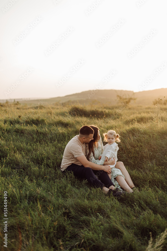 Beautiful caucasian family of father, mother and little daughter posing on camera on background of green field