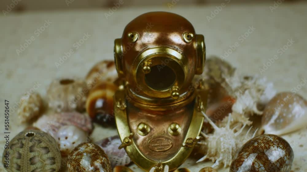 A copy of an old copper diving helmet surrounded by beautiful multi-colored sea shells against the background of the table surface imitating sea sand. Marine romance concept A copy of an old copper diving helmet surrounded by beautiful multi-colored sea shells against the background of the table surface imitating sea sand. Marine romance concept