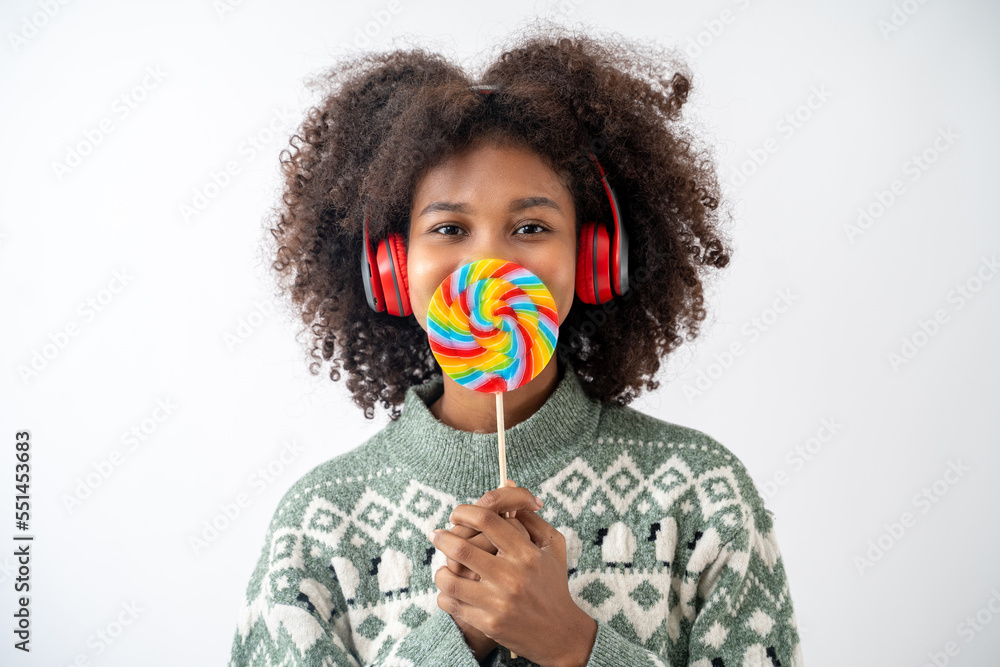 Portrait of young attractive african american woman with curly hair and headphone holding colorful lollipop in studio on white background.