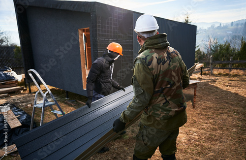 Male builders installing black corrugated iron sheet used as facade of future cottage. Men workers building wooden frame house. Carpentry and construction concept. © anatoliy_gleb
