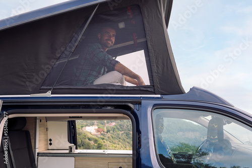 young man looking out over the landscape sitting in the roof tent of his camper van, concept of nature travel and nomadic lifestyle
