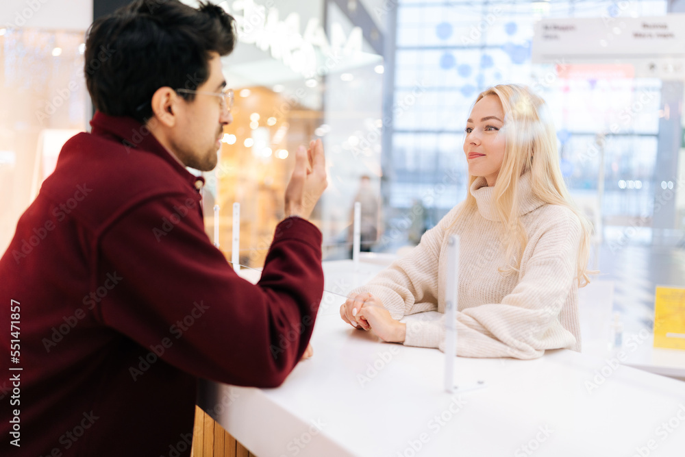 Back view from shoulder of handsome man chatting through glass partition with charming young blonde woman standing in bright hall of shopping mall. Concept of lifestyle social distancing and work. Back view from shoulder of handsome man chatting through glass partition with charming young blonde woman standing in bright hall of shopping mall. Concept of lifestyle social distancing and work.